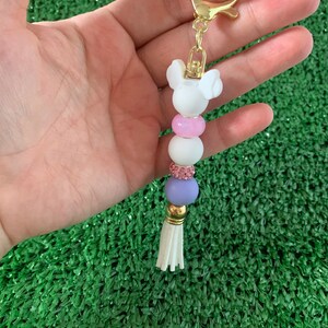 May include: A white and pink beaded keychain with a white Mickey Mouse charm and a white tassel. The keychain is attached to a gold key ring.