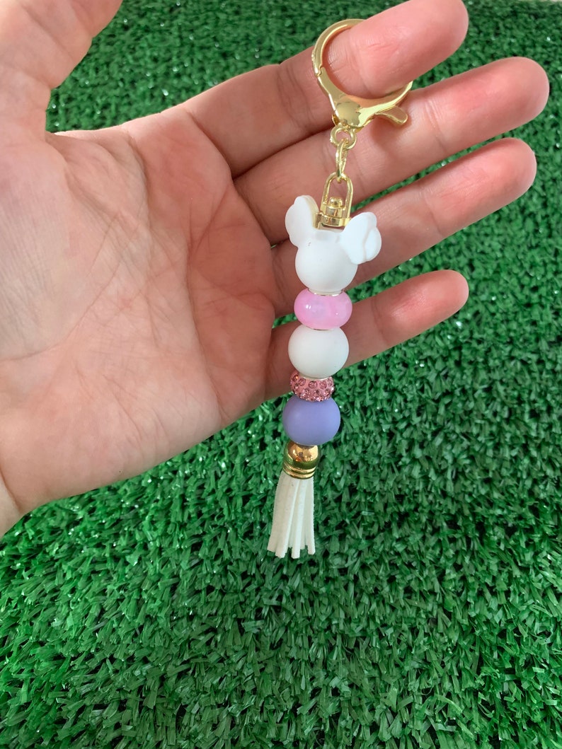May include: A white and pink beaded keychain with a white Mickey Mouse charm and a white tassel. The keychain is attached to a gold key ring.