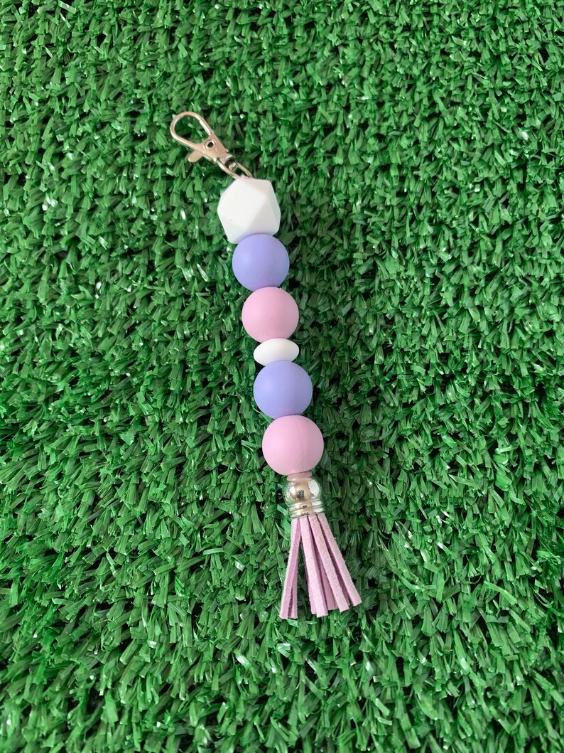 May include: A purple, pink, and white beaded keychain with a tassel. The keychain has a silver clasp.