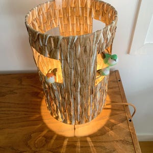 Puede incluir: Una lámpara caprichosa con forma de tronco de árbol, hecha de fibras naturales tejidas. La lámpara presenta ventanas recortadas con pequeñas figuras de pájaros de colores. Una pequeña figura de pájaro se encuentra en la parte superior. La lámpara emite un brillo cálido.