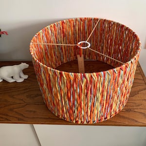 Peut inclure: Un abat-jour rond en tissu tissé dans des tons d'orange, rouge, jaune et bleu. L'abat-jour est doté d'une simple bague blanche en haut et d'une étiquette brune sur laquelle est inscrit "Atelier Lisa".