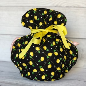 May include: A quilted teapot cozy with a black background and a pattern of yellow lemons and green leaves. The cozy is tied with a yellow ribbon. The teapot itself is a light pink color.