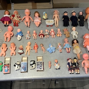 Puede incluir: Una colección de muñecas vintage, algunas con caras pintadas y otras con cuerpos de tela, en varias poses y atuendos. Las muñecas están dispuestas sobre una mesa con una superficie blanca. Algunas de las muñecas sostienen accesorios, como un bolso, un libro o una flor.