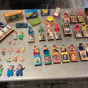 Könnte beinhalten: Eine Sammlung von Vintage-Spielzeugen, darunter Puppen, Figuren in Streichholzschachteln und Miniaturzubehör wie eine Schaukel und ein Auto. Die Puppen sind in verschiedenen Outfits und Farben gekleidet, einige werden in Streichholzschachteln präsentiert.