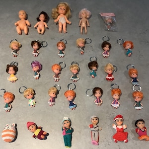 May include: A collection of vintage dolls and doll keychains. The dolls vary in size and style, with some featuring blonde or brown hair. The keychains have small doll figures with various outfits. There are also several small, naked dolls and a small bag of doll accessories.