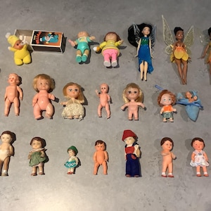 Può includere: Una collezione di bambole e figurine vintage, tra cui bambole per bambini, fate con le ali e una piccola bambola con un cappello giallo. Le bambole sono in varie dimensioni e abiti, disposte su una superficie grigia.