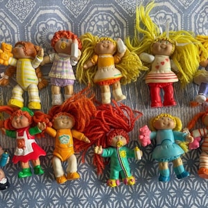Puede incluir: Una colección de muñecas Cabbage Patch Kids vintage, cada una con atuendos y peinados únicos. Las muñecas presentan varios colores, incluyendo amarillo, naranja, rosa y azul, y están dispuestas sobre una superficie azul estampada.