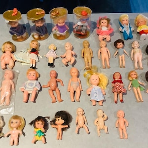 Peut inclure: Une collection de trente-cinq petites poupées en plastique disposées en rangées. Certaines poupées sont nues, tandis que d'autres portent diverses tenues, notamment des robes, des chemises et des chapeaux. Quatre poupées sont dans des tubes.