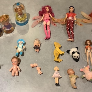 Puede incluir: Una colección de muñecas y figuras vintage, que incluye muñecas de hadas con alas, un panda, un cerdo y figuras en miniatura en recipientes de vidrio. Las muñecas varían en tamaño y estilo, con colores brillantes.
