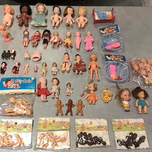 Könnte beinhalten: Eine Sammlung von Vintage-Babypuppen in verschiedenen Posen und Outfits. Die Puppen sind aus Plastik und haben bemalte Gesichtszüge. Einige Puppen tragen Kleider, andere Hosen oder Röcke. Es gibt auch einige Puppen, die nackt sind. Die Puppen sind auf einer weißen Oberfläche angeordnet.