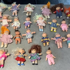 Puede incluir: Una colección de muñecas Cabbage Patch Kids vintage, cada una con peinados y atuendos únicos. Las muñecas están en varias poses, mostrando sus coloridos vestidos, monos y accesorios. Las muñecas están dispuestas sobre una superficie gris.