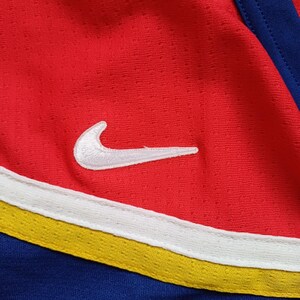 Op de afbeelding: Close-up van een rood, wit en blauw sport shirt met een wit Nike swoosh logo. Het shirt heeft een gele streep.