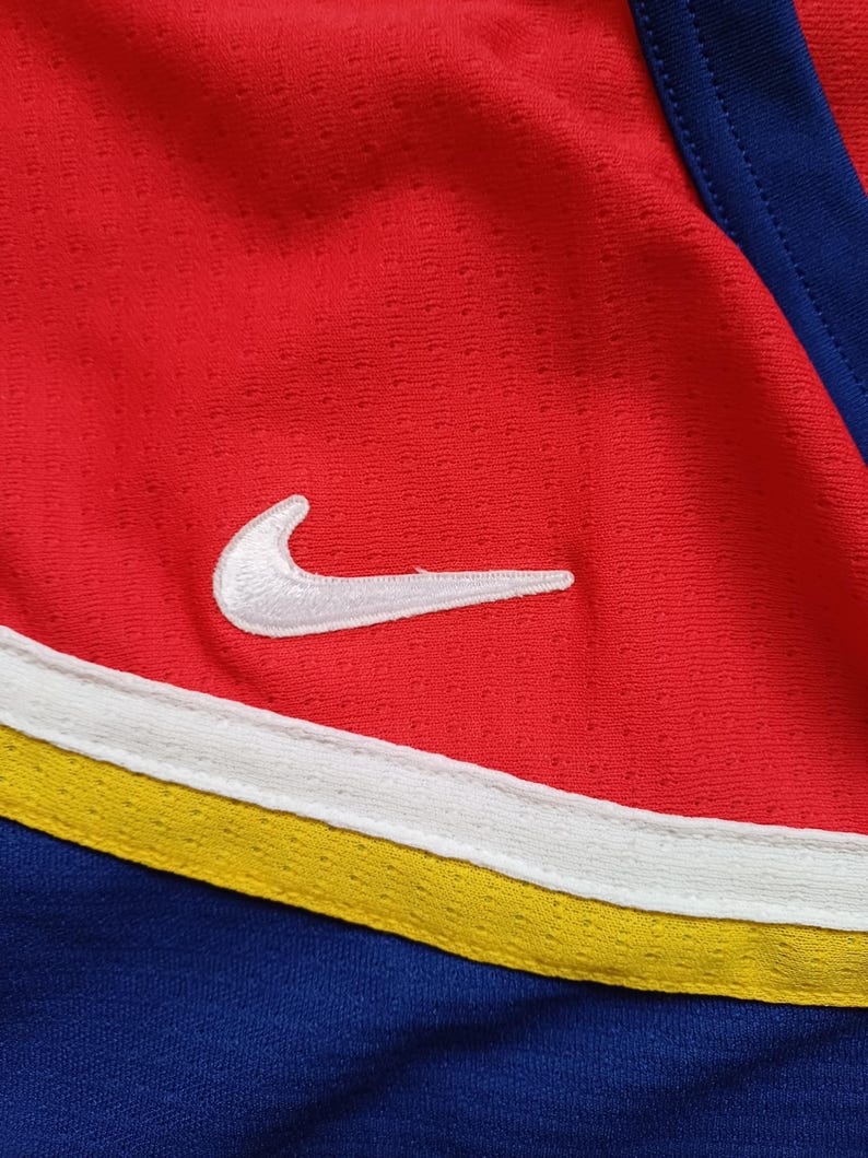 Op de afbeelding: Close-up van een rood, wit en blauw sport shirt met een wit Nike swoosh logo. Het shirt heeft een gele streep.