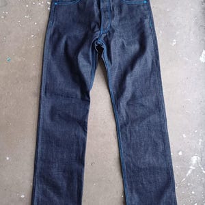 Könnte beinhalten: Dunkelblaue Jeans aus Denim mit blauen Nähten und einem Knopfverschluss. Die Jeans hat einen geraden Beinverlauf und einen rohen Saum. Der Markenname "WESC" ist sichtbar.