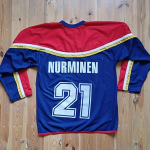 Op de afbeelding: Een blauw hockeytrui met gele en rode bies. De achterkant van de trui heeft de naam "NURMINEN" en het nummer "21" in wit.