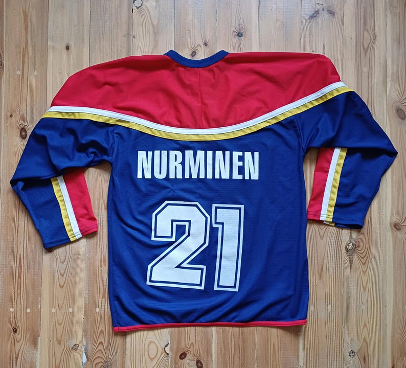 Op de afbeelding: Een blauw hockeytrui met gele en rode bies. De achterkant van de trui heeft de naam "NURMINEN" en het nummer "21" in wit.