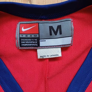 Op de afbeelding: Een rood en donkerblauw sporttrui met een wit label waarop staat "TEAM ENGINEERED TO THE EXACT SPECIFICATIONS OF CHAMPIONSHIP ATHLETES." Het label heeft ook een wit label met de tekst "NAME/#" en een grijs label met de tekst "M" en het Nike swoosh-logo. Het label vermeldt ook "MADE IN CANADA".