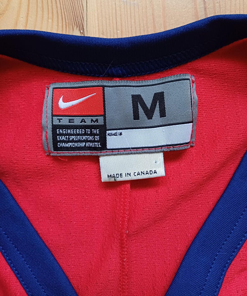 Op de afbeelding: Een rood en donkerblauw sporttrui met een wit label waarop staat "TEAM ENGINEERED TO THE EXACT SPECIFICATIONS OF CHAMPIONSHIP ATHLETES." Het label heeft ook een wit label met de tekst "NAME/#" en een grijs label met de tekst "M" en het Nike swoosh-logo. Het label vermeldt ook "MADE IN CANADA".