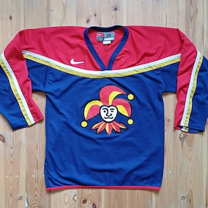 Op de afbeelding: Een blauw, rood en geel hockeytrui met een narrenlogo op de voorkant. De trui heeft een Nike-logo op de mouw en de maat "M" op het label.