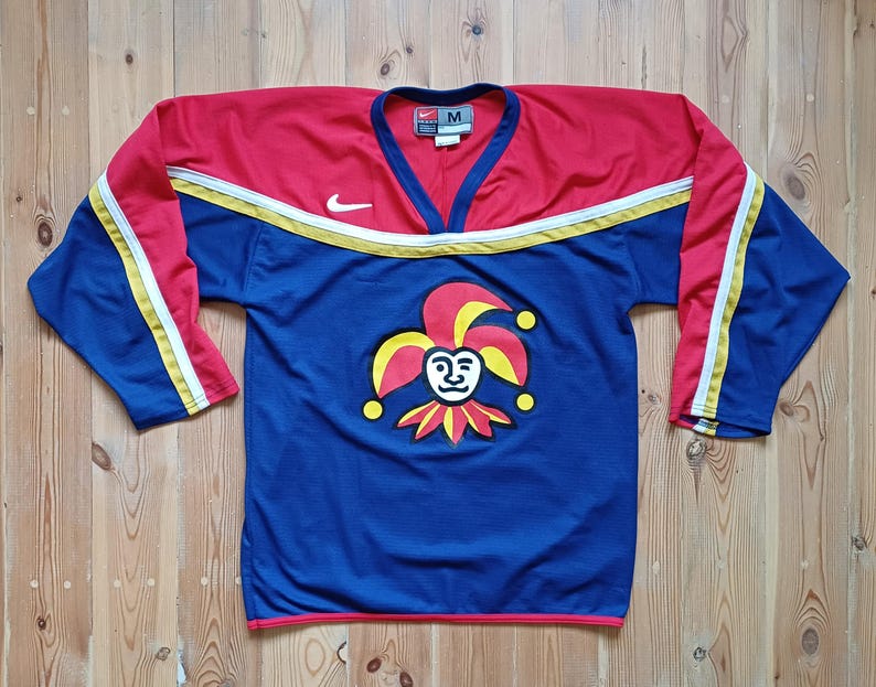 Op de afbeelding: Een blauw, rood en geel hockeytrui met een narrenlogo op de voorkant. De trui heeft een Nike-logo op de mouw en de maat "M" op het label.