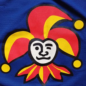 Op de afbeelding: Een blauwe stoffen patch met een rode, gele en zwarte narrenmuts en gezicht. De narrenmuts heeft drie punten en het gezicht heeft een witte achtergrond met zwarte ogen, neus en mond.