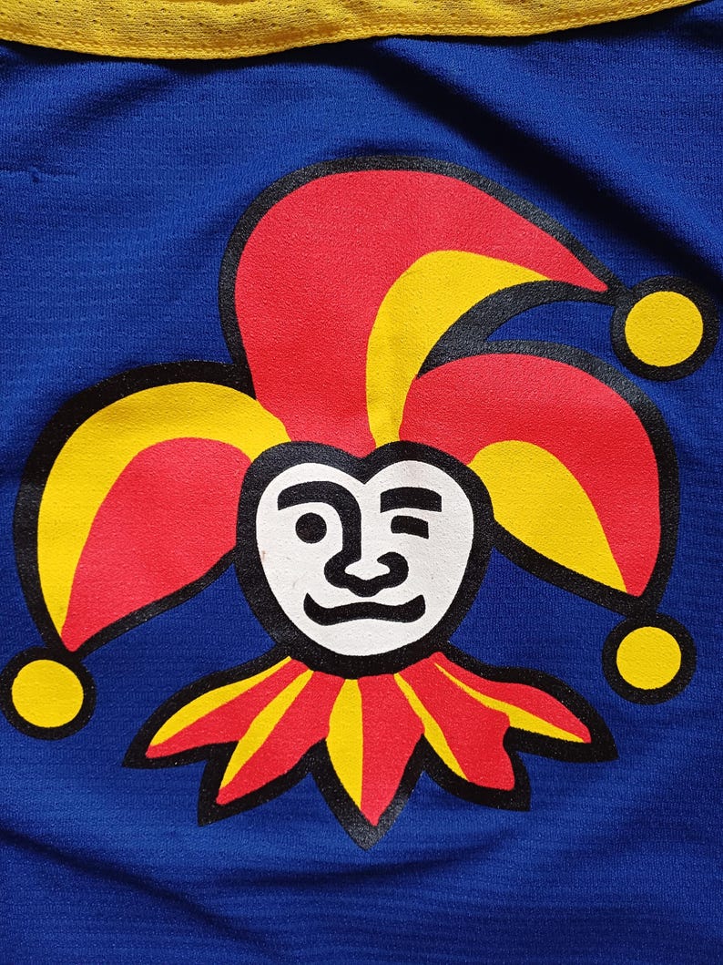 Op de afbeelding: Een blauwe stoffen patch met een rode, gele en zwarte narrenmuts en gezicht. De narrenmuts heeft drie punten en het gezicht heeft een witte achtergrond met zwarte ogen, neus en mond.