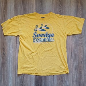 Op de afbeelding: Geel T-shirt met korte mouwen met een blauwe en witte afbeelding van handbalspelers en de tekst "Sverige Handboll".