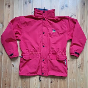 Puede incluir: Una chaqueta impermeable roja con una cremallera negra y un pequeño logotipo en el pecho izquierdo. La chaqueta tiene dos bolsillos grandes en la parte delantera y un cordón en la cintura.