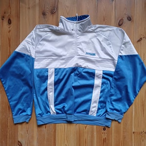 Op de afbeelding: Een blauw en wit trainingsjack met een witte rits en een "DIADORA" logo op de borst.