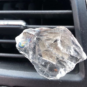 May include: A clear quartz crystal, with a slightly cloudy appearance, is placed in a car's air vent.