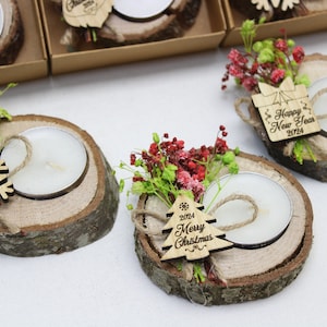 May include: Set of three wooden candle holders with small white candles. Each holder is decorated with a small sprig of red and green flowers and a wooden tag with a holiday message. The tags read "Happy New Year 2024", "Happy New Year 2024", and "2024 Merry Christmas".