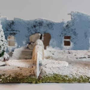 May include: A miniature diorama showing a snowy scene with a damaged blue building, two snow-covered fir trees, and a small aeroplane. The base is green with white snow.