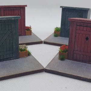 May include: Four miniature wooden sheds in red and green, each with a small flower box and a metal bin. The sheds are set on grey platforms, creating a small-scale street scene. The scene includes faux grass.
