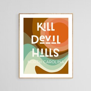 May include: Framed art print featuring the text "KILL DEVIL HILLS NORTH CAROLINA" in white, set against a retro-style background of layered brown, orange, and teal shapes. The artwork has a minimalist design.