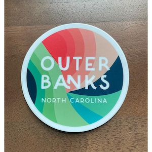Puede incluir: Una pegatina circular blanca con un dise&ntilde;o abstracto colorido. El texto "Outer Banks" est&aacute; en letras blancas sobre un fondo azul. El texto "North Carolina" est&aacute; en letras blancas sobre un fondo verde.