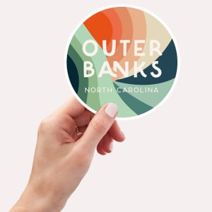 Puede incluir: Una pegatina redonda con un dise&ntilde;o abstracto colorido en tonos naranja, verde y azul. La pegatina dice "OUTER BANKS NORTH CAROLINA" en texto blanco.