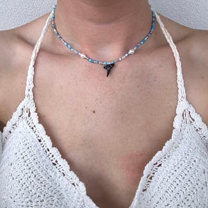 May include: A delicate necklace featuring a shark tooth pendant. The necklace is made of small blue, white and silver beads. The shark tooth is black and has a pointed tip.