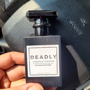 Puede incluir: Una botella de colonia indígena Deadly de vidrio negro con aroma a hierba dulce y cedro. La botella tiene una etiqueta negra con texto blanco que dice "Deadly indigenous COLOGNE Sweetgrass&Cedar www.niikicraft.ca".