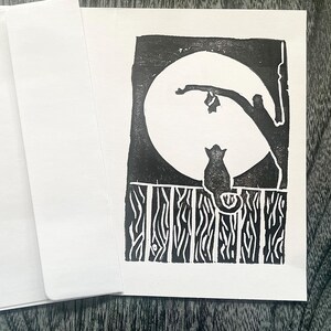 May include: A black and white linocut print depicting a cat on a fence, with a full moon in the background. The print is on white paper, with a white envelope and card. The artwork has a minimalist, artistic style.