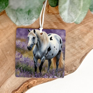 May include: Square air freshener featuring a white horse with black spots, standing in a field of purple flowers. The horse has a flowing white mane and tail. The air freshener is hanging by a white string.