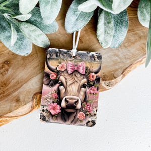 Pode incluir: Um ambientador retangular com uma vaca adornada com um laço rosa e flores. A vaca tem pelo castanho e branco, com flores rosa e vermelhas. O fundo é uma mistura de rosa e preto.