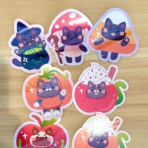 May include: A collection of colorful stickers featuring cartoon cats in Halloween-themed costumes. The stickers include a witch cat, a cat in a mushroom costume, a cat in a candy corn costume, and a cat in a pumpkin costume.