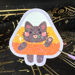 May include: A holographic sticker featuring a cartoon cat inside a candy corn shape. The cat is black with pink cheeks and a smiling face. The candy corn has white, yellow, and orange sections with star-shaped sparkles.