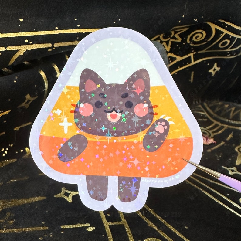 May include: A holographic sticker featuring a cartoon cat inside a candy corn shape. The cat is black with pink cheeks and a smiling face. The candy corn has white, yellow, and orange sections with star-shaped sparkles.