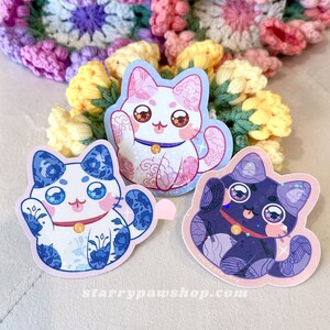 May include: Three cat-shaped stickers with a waving paw design. The stickers are in blue and white, pink and white, and purple and pink. Each cat has a gold bell collar and large, expressive eyes. The website address "starrypawshop.com" is printed on one sticker.