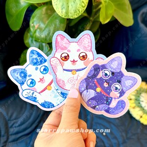 May include: Three cat-shaped stickers in blue, pink, and purple. The blue cat has a floral design, the pink cat is waving, and the purple cat has a starry pattern. Each sticker has large eyes and a small collar.
