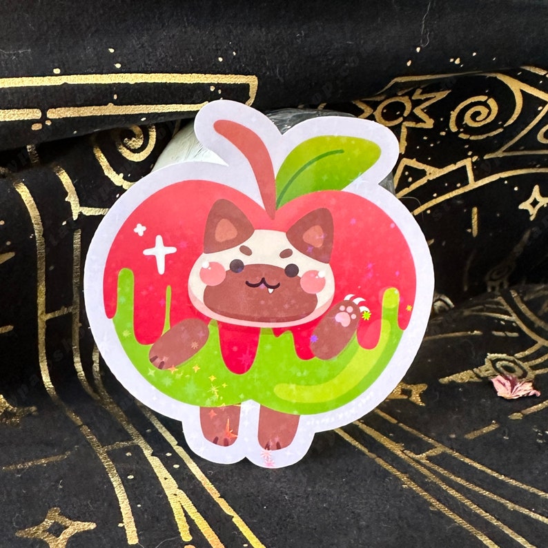 May include: A sticker featuring a cartoon cat inside a red apple with green dripping "juice". The cat has brown and white fur, with a small candy cane in its paw. The sticker has a glittery finish.