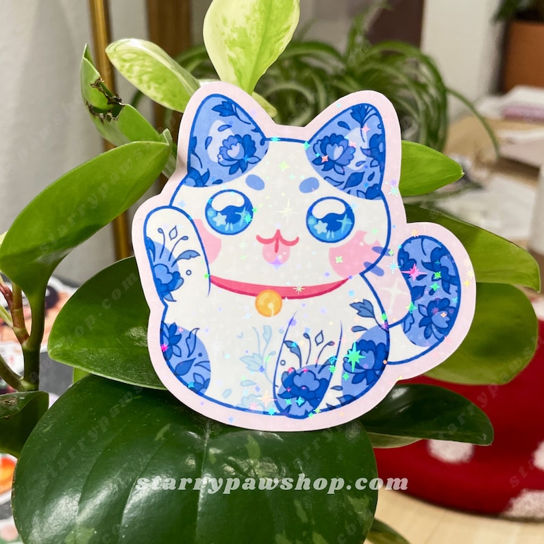 May include: A die-cut sticker of a lucky cat with blue floral patterns on a white background. The cat has large blue eyes, pink cheeks, and a pink collar with a gold bell. The sticker has a holographic finish and the text "starrypawshop.com".