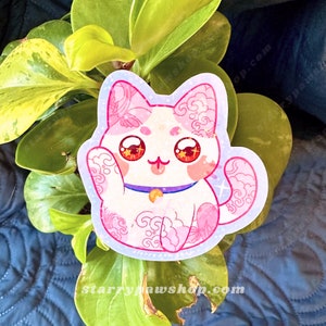 May include: A sticker featuring a white cat with pink accents and floral patterns. The cat has large, red eyes and a purple collar with a gold bell. The sticker is surrounded by a light blue border with star accents.