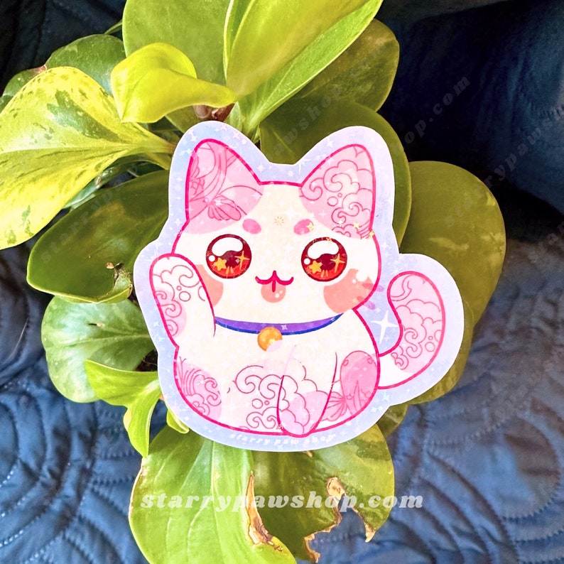 May include: A sticker featuring a white cat with pink accents and floral patterns. The cat has large, red eyes and a purple collar with a gold bell. The sticker is surrounded by a light blue border with star accents.